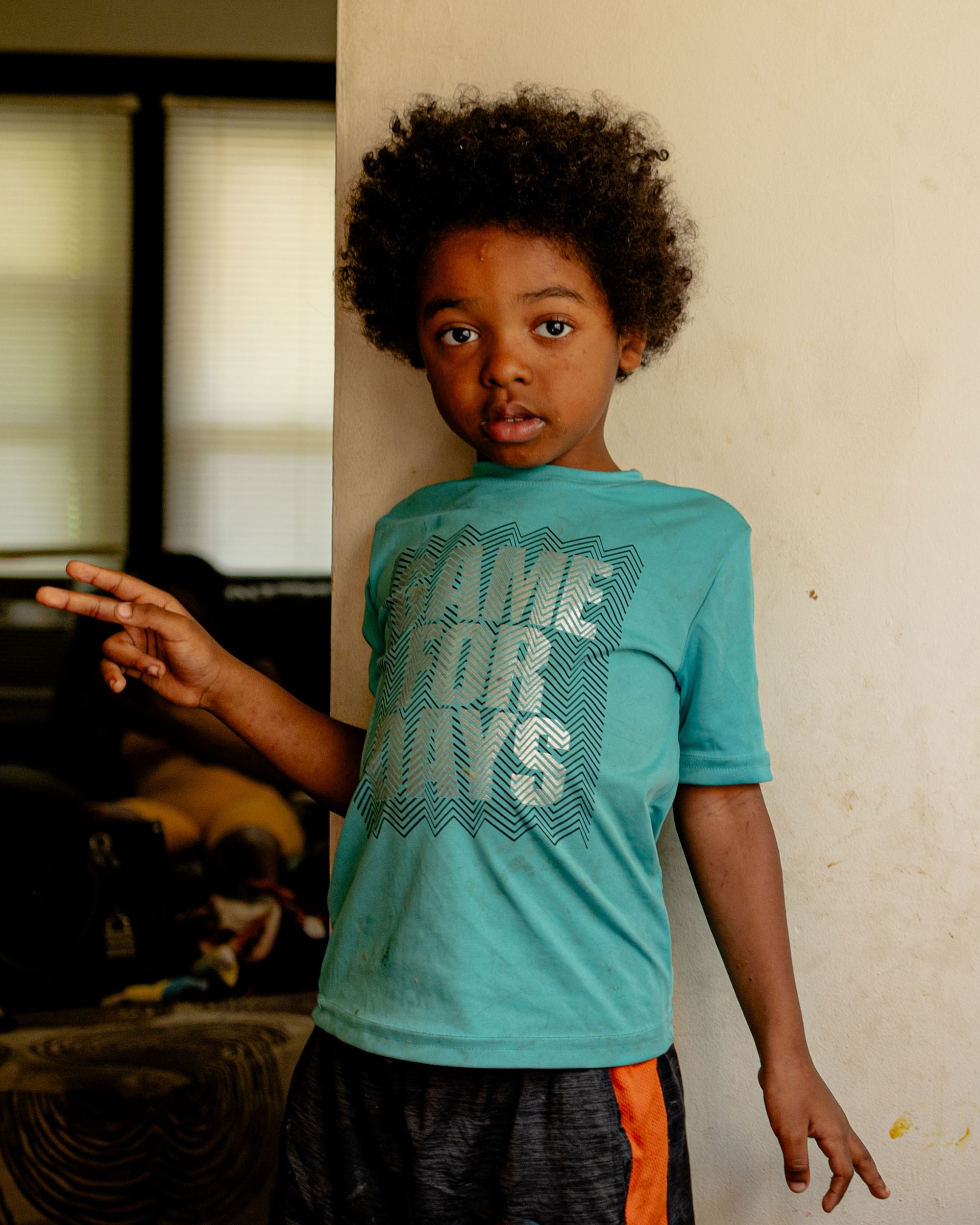 A photo of a Black boy with a turquoise shirt looking into the camera while leaning against a wall. 