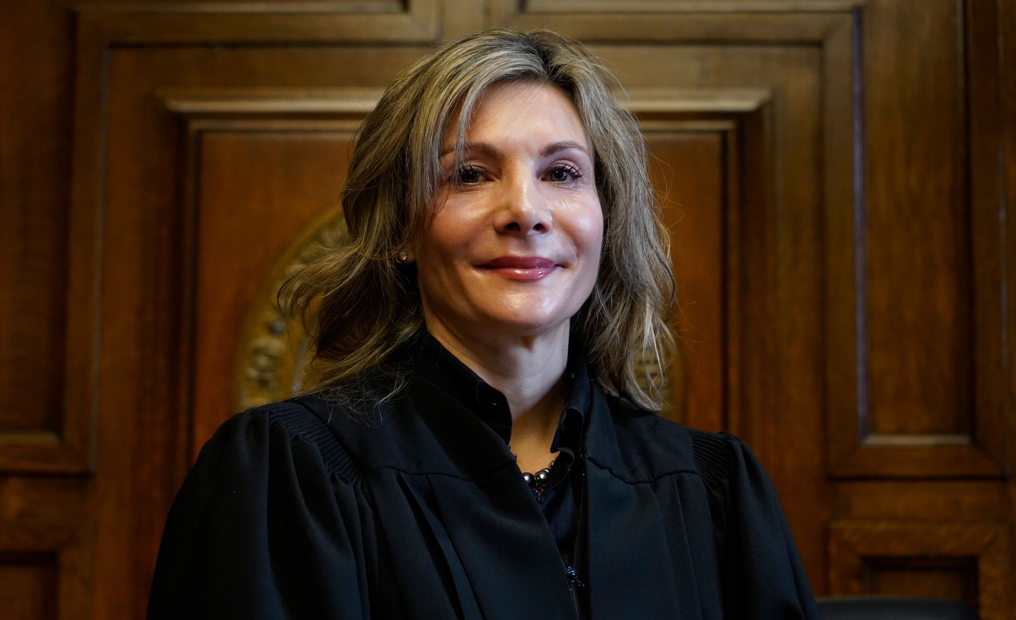 A White, blonde woman stands in a black robe behind a judge’s podium. 