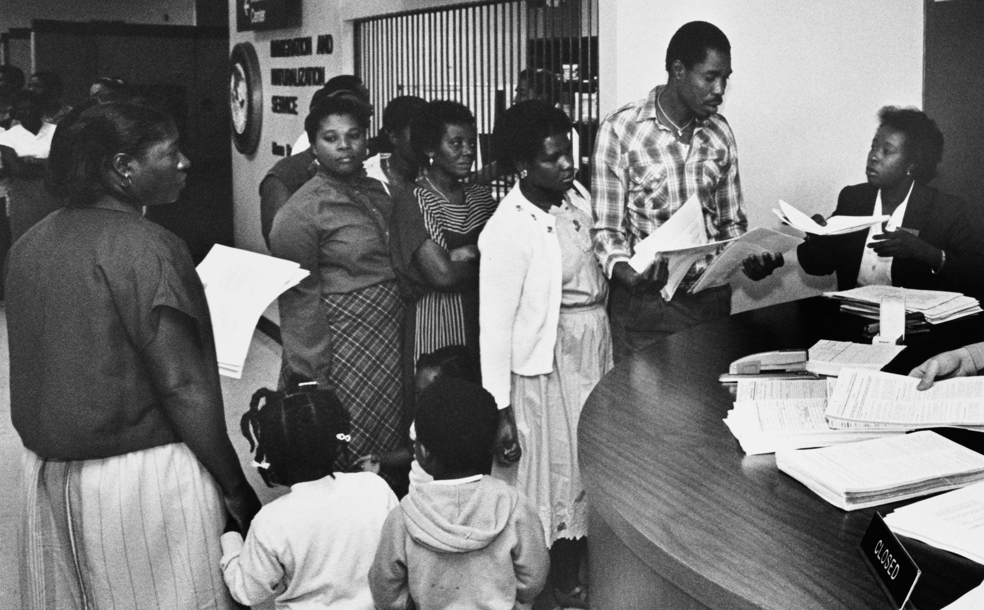 A black-and-white photo shows a Black woman holding a folder speaking to a Black man holding paperwork in front of a desk with piles of forms. A group of Black women and children stand nearby. A sign for the "Immigration & Naturalization Service" is in the background, with the agency's logo. 
