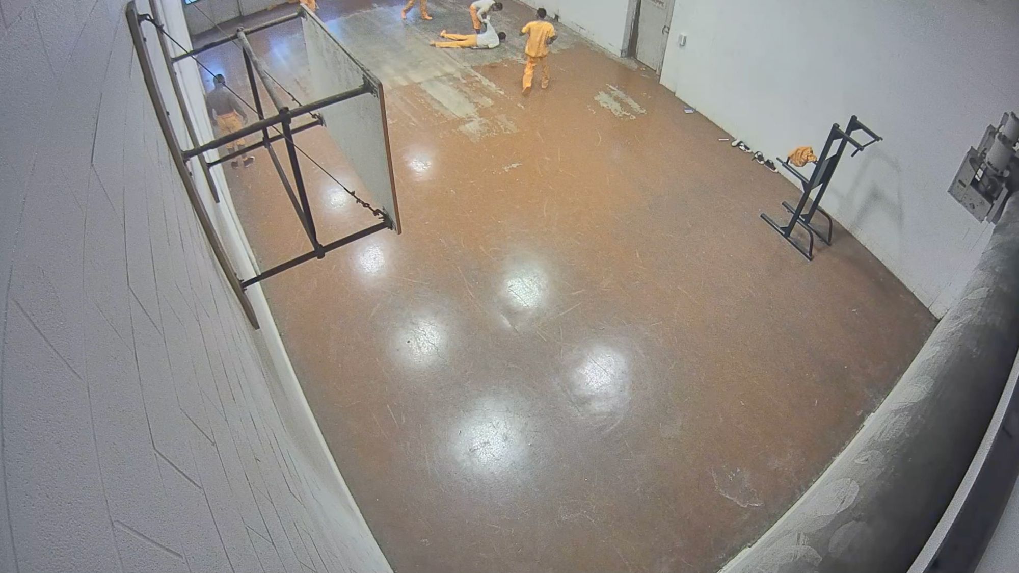 Three still images from a surveillance camera show an indoor gym at a jail. In the first frame, a man in an orange and white jail uniform is on the ground as other men approach him. In the second frame, a correctional officer kneels over the man on the floor. In the third frame, a group of correctional officers surround the man on the floor.