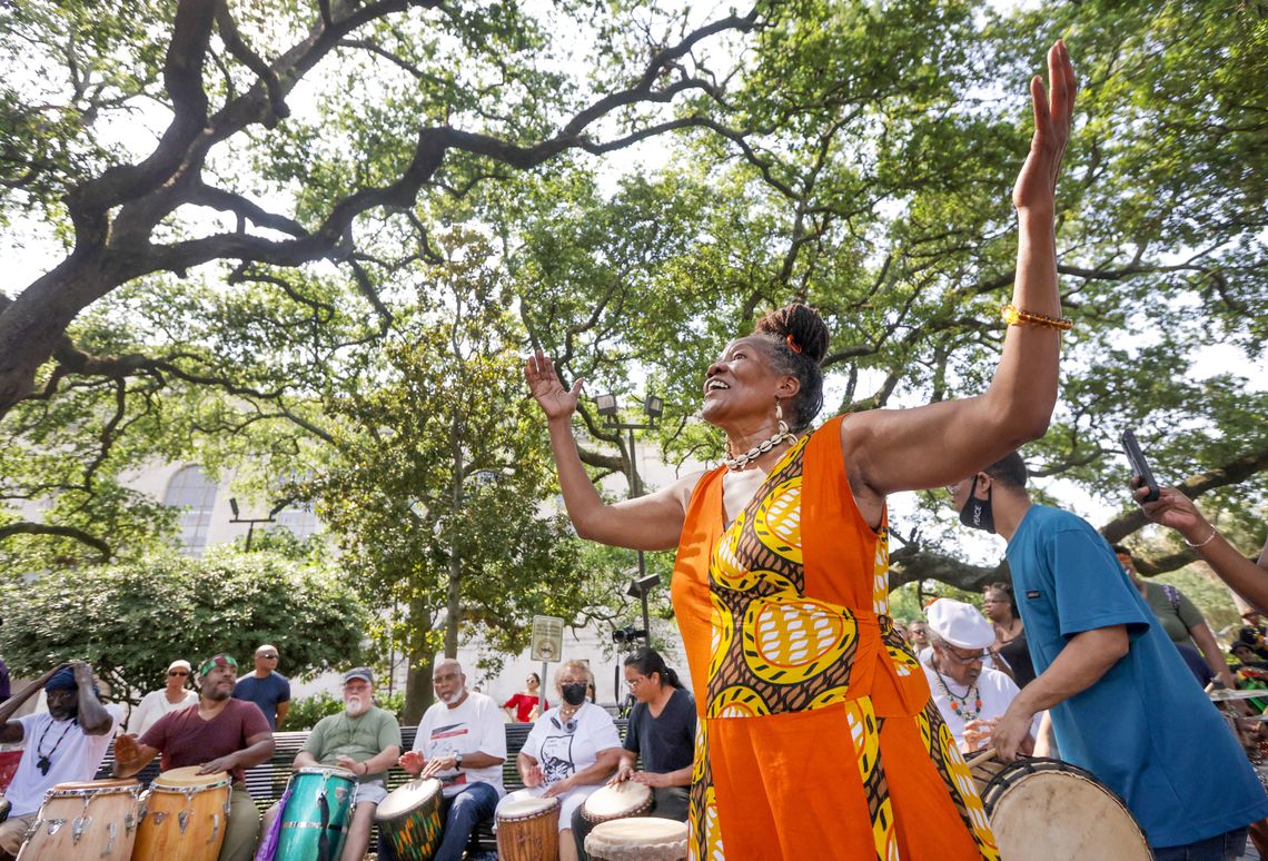 A 69-year-old Black woman wearing an orange-yellow dress and a white seashell necklace raises her arms in the air during an outside gathering. Individuals playing various drums are seen in the background.