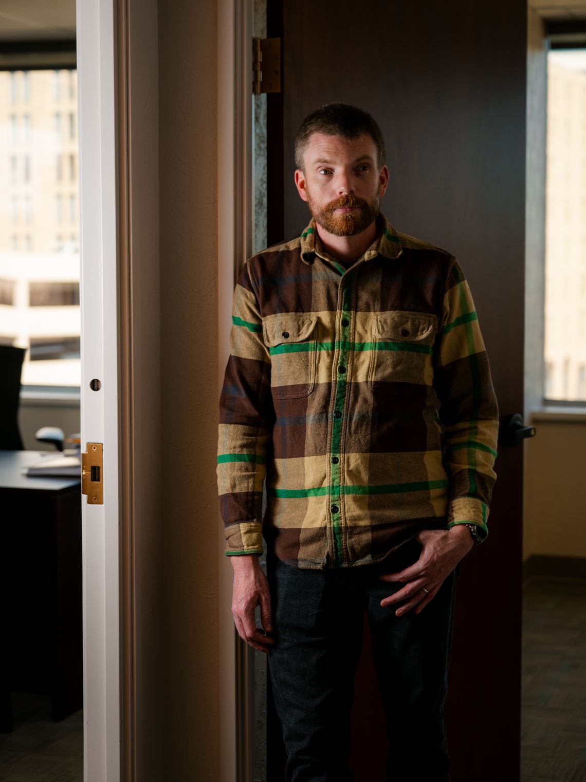 A photo shows a White man with brown hair and a beard. He is wearing a brown, yellow and green plaid shirt and is standing between two doorways. 