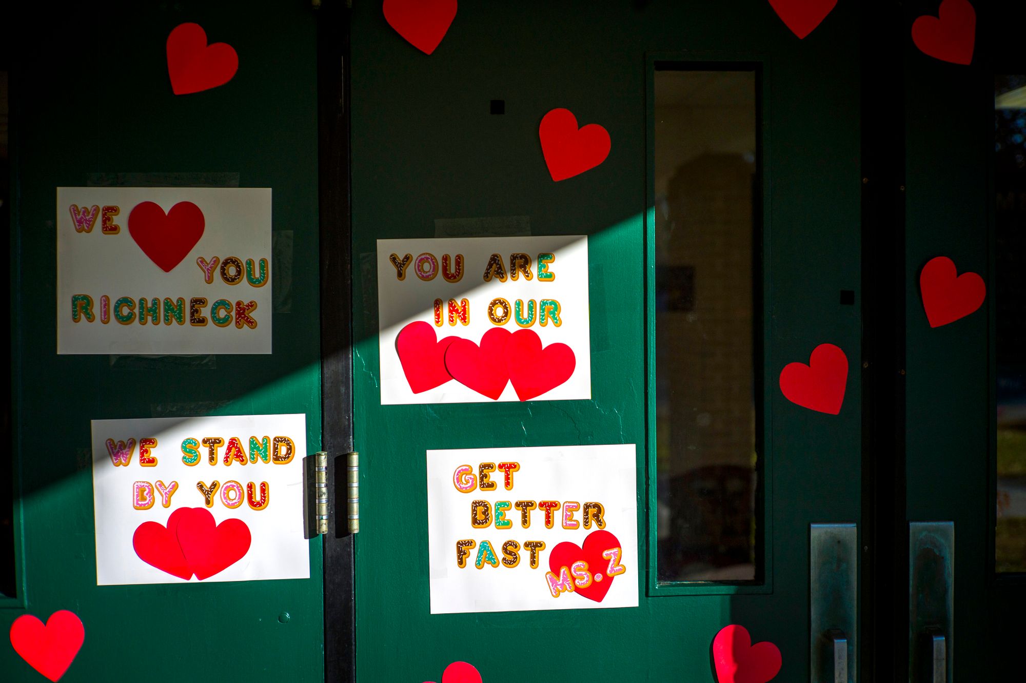 Signs and red paper hearts are taped to a green school door. The signs read as messages of support, such as: "We love you Richneck," "You are in our hearts," "We stand by you," and "Get better fast Ms. Z." 