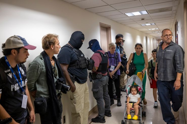 A photo shows a woman in a green dress pushing a stroller with a toddler down a hall. Federal agents, some of whom are masked, stand along the side of the hallway.  