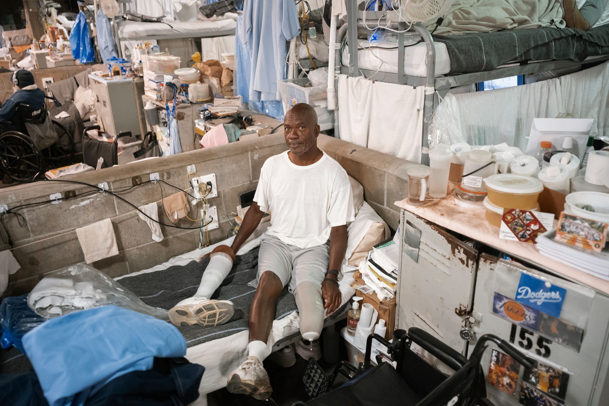 A photo shows a Black man sitting in his living space, which has a bed, which is among many beds in the open space. There is a cabinet with a postcard or stickers with the Dodger’s logo on it and another with the Laker’s logo on it. There are tupperwares on top of the cabinet, which also serves as a desk. 