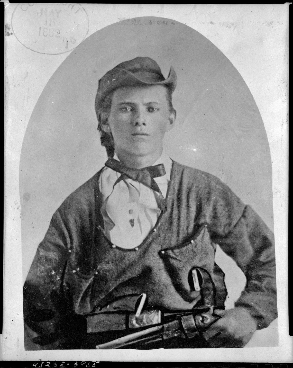 A black-and-white photo shows a White man wearing a hat and holding a gun in his left hand. 