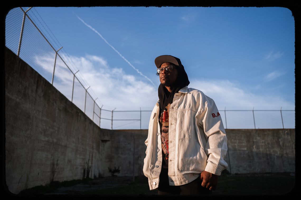 A photo shows Minister King X, a Black man with glasses, wearing a baseball cap and a white jacket, standing outdoors, surrounded on two sides by concrete walls with a chain-link fence on top. 