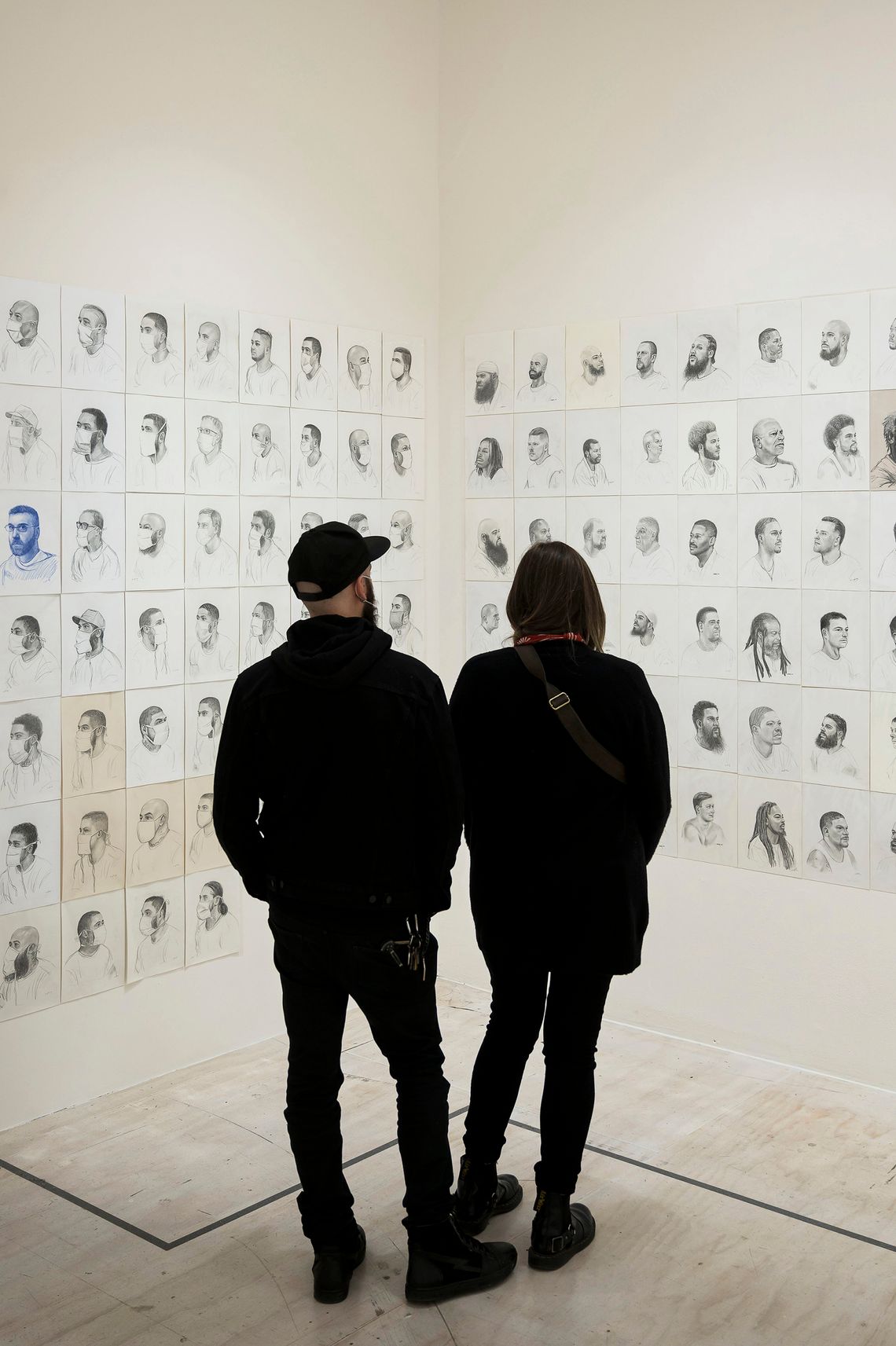 Two people, wearing all black clothing, stand and view more than a hundred portraits of incarcerated men displayed on an art gallery wall. 