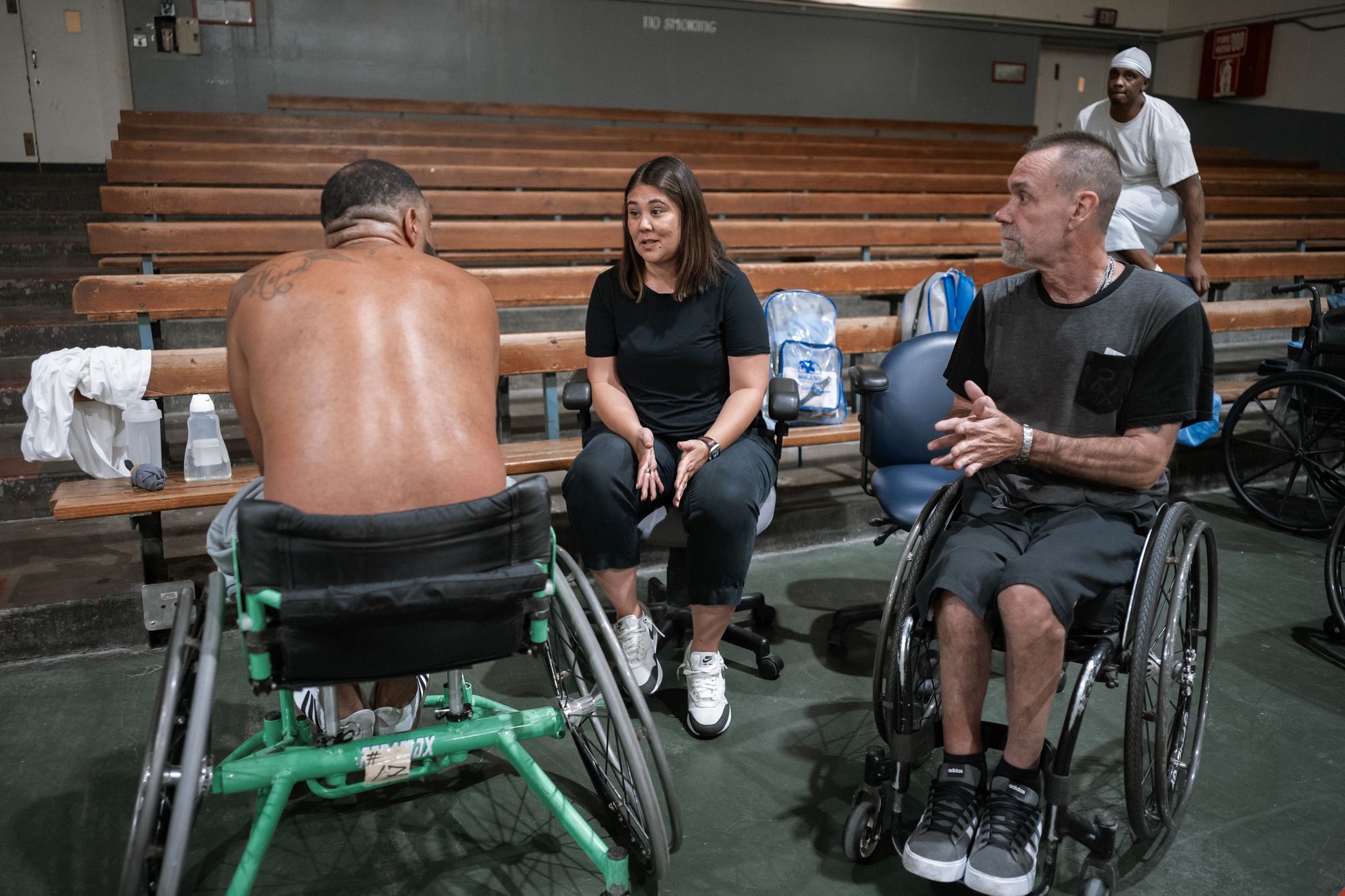 A photo shows Coach Mieko Chambers, a woman with light-toned skin and dark brown hair, talking to a man with medium-toned skin sitting in a wheelchair. Another man with light-toned skin, in a wheelchair sits nearby. 