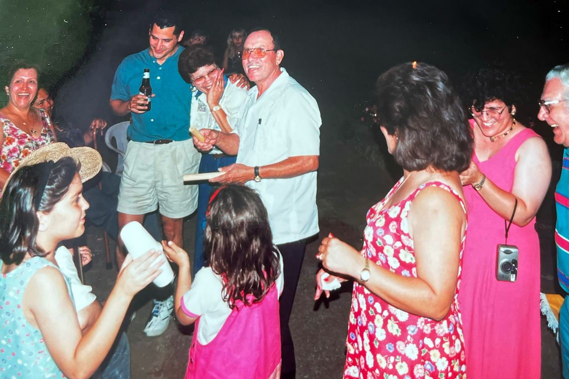 A middle-aged man wearing a white shirt and sunglasses laughs with family members during a party, in a photo from the 1980s. 