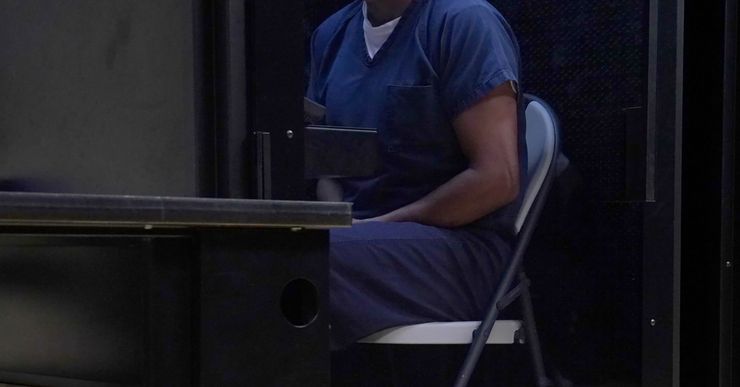 A man wearing a blue uniform sits on a metal folding chair in a small room. 