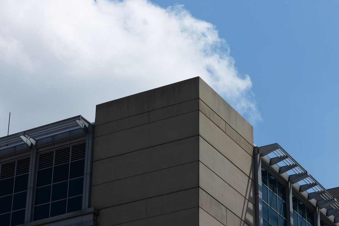 A wide photo shows the outside corner of a jail with concrete walls and blue windows. One side of the building is in harsh light while the other is shaded. A cloudy blue sky is visible above the building. 