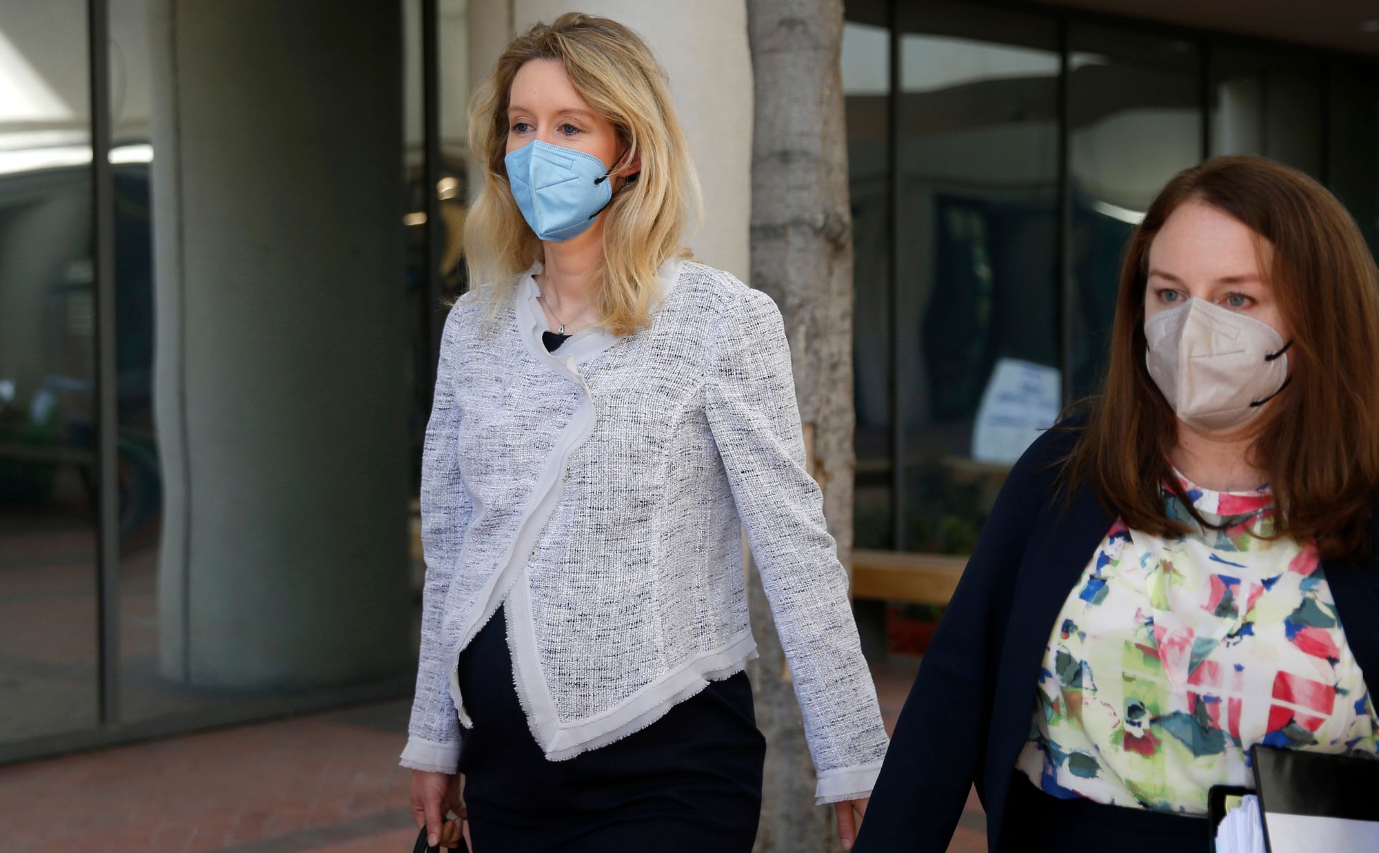 Elizabeth Holmes, a White woman with blonde hair, is visibly pregnant and walking with a female member of her defense team in a photo.  