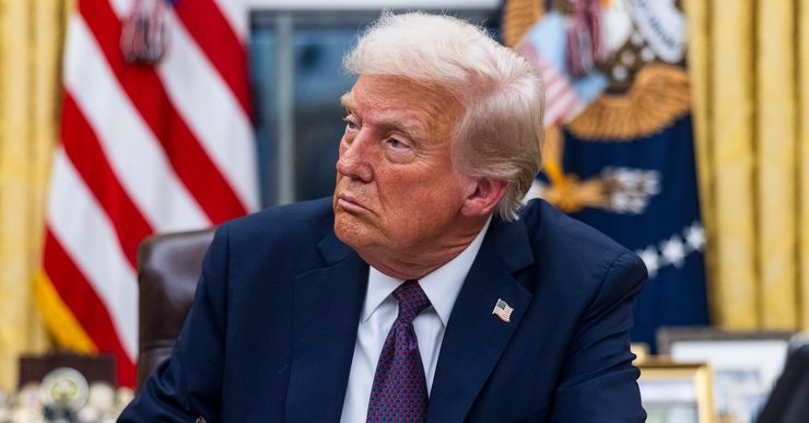 Trump, a White man in a navy suit and a red and navy tie, signs a document while sitting in the Oval Office. 