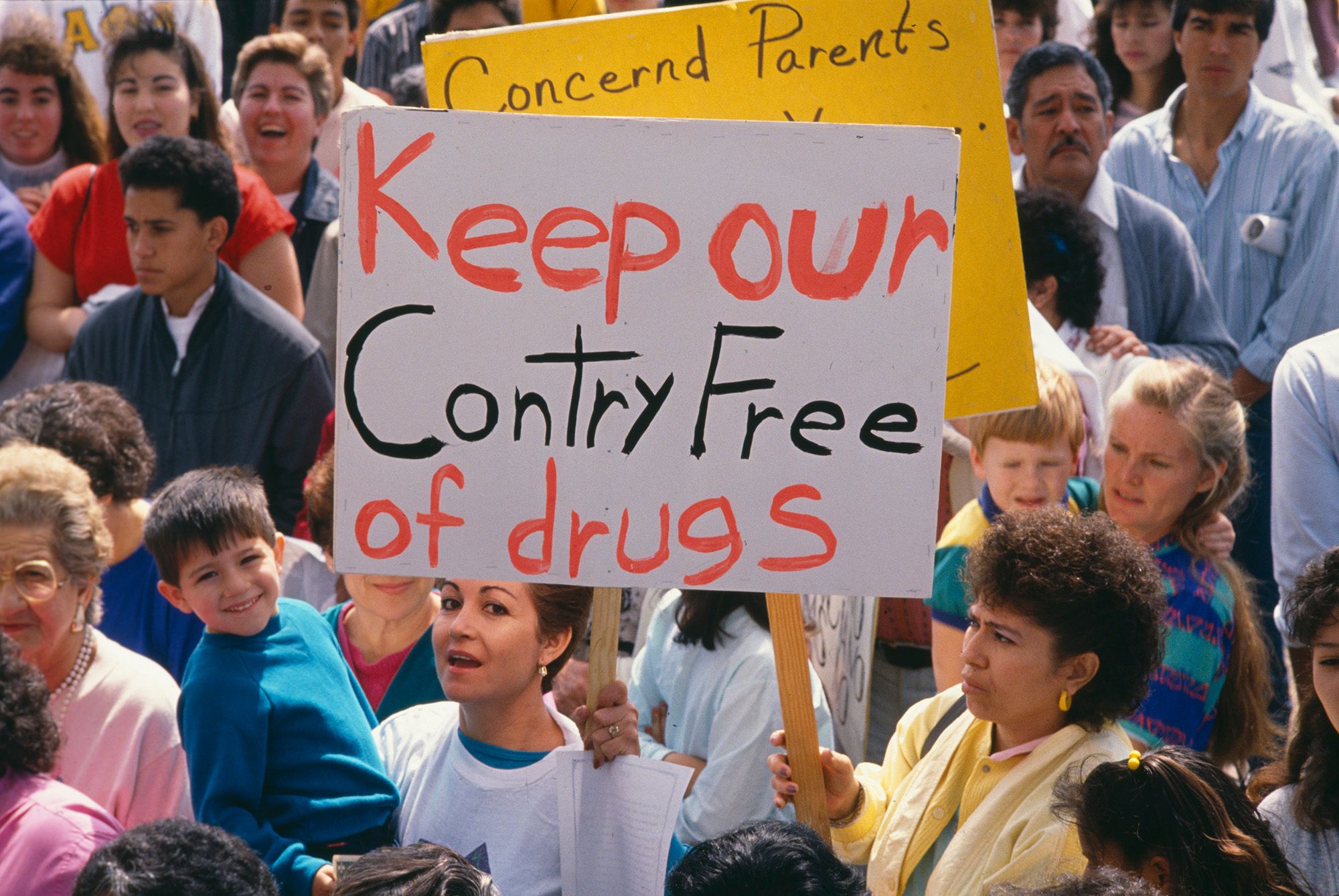 A photo shows a large crowd of demonstrators. One person is holding a sign that reads, "Keep our country free of drugs."  