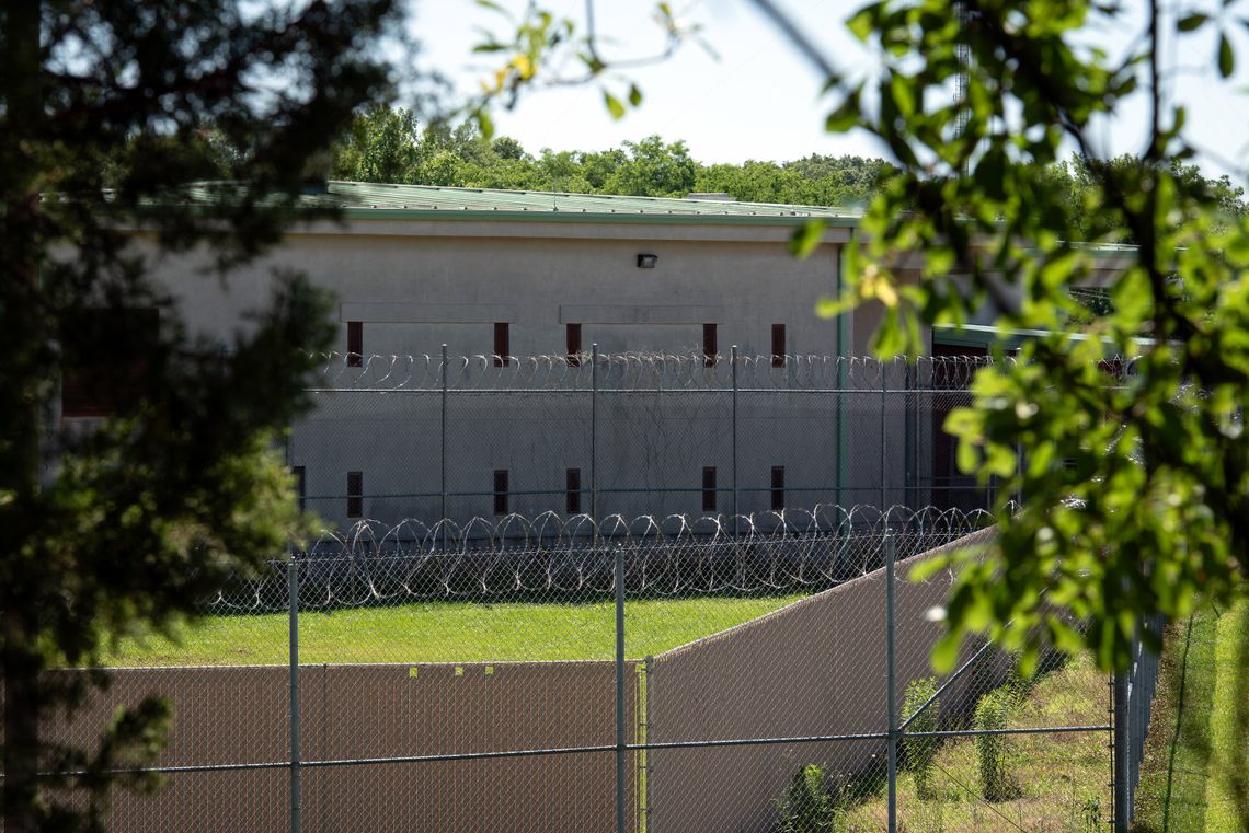 A photo of a building behind a barbed wire fence. The building has a light green roof and two levels of narrow windows. In the foreground, leaves and branches from a tree are out of focus.