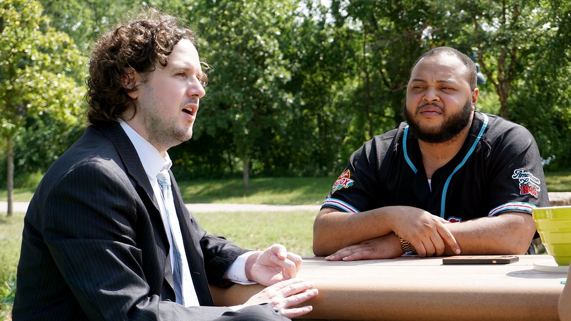 A Black man, wearing a black baseball jersey, listens to a White male attorney speaking while sitting at a picnic table. 