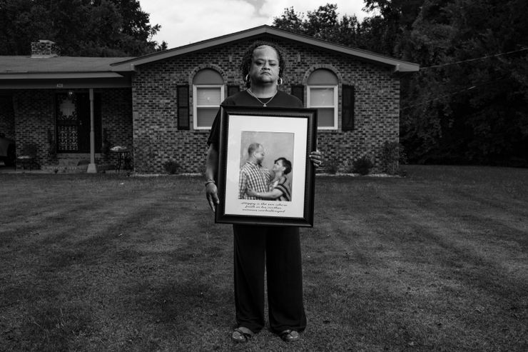 A photo shows a Black woman with curly hair in a ponytail holding a framed picture while posing for a portrait in front of her home. The framed picture shows her embracing and looking toward her son, a Black man with a plaid shirt and glasses, in front of a green background.
