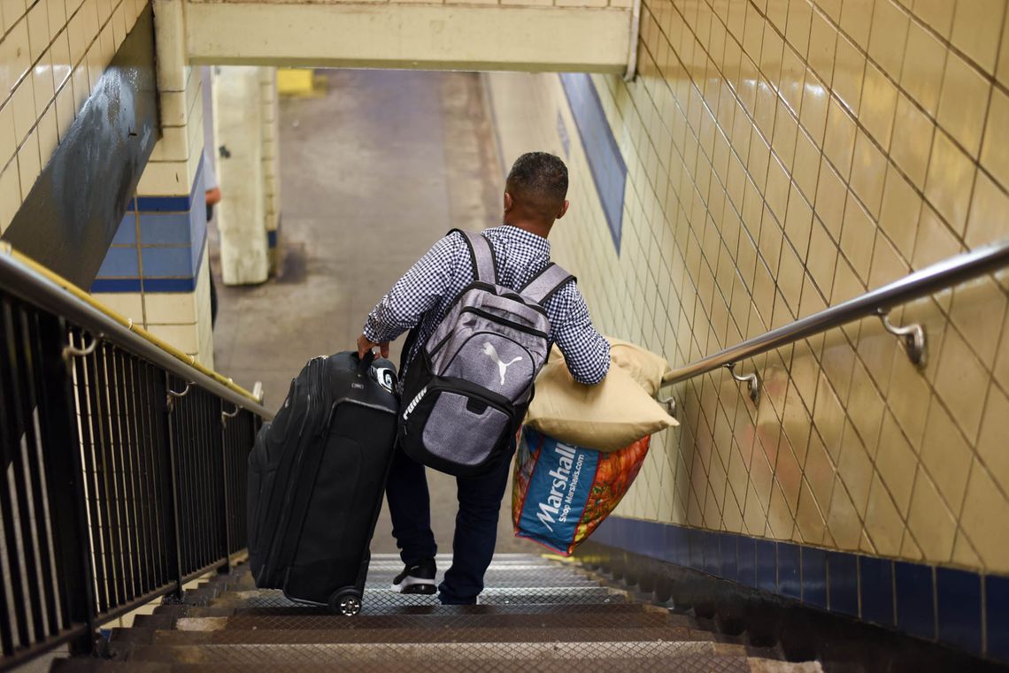 A man in a blue-checkered shirt descends a subway staircase lugging a large suitcase and a shopping bag.