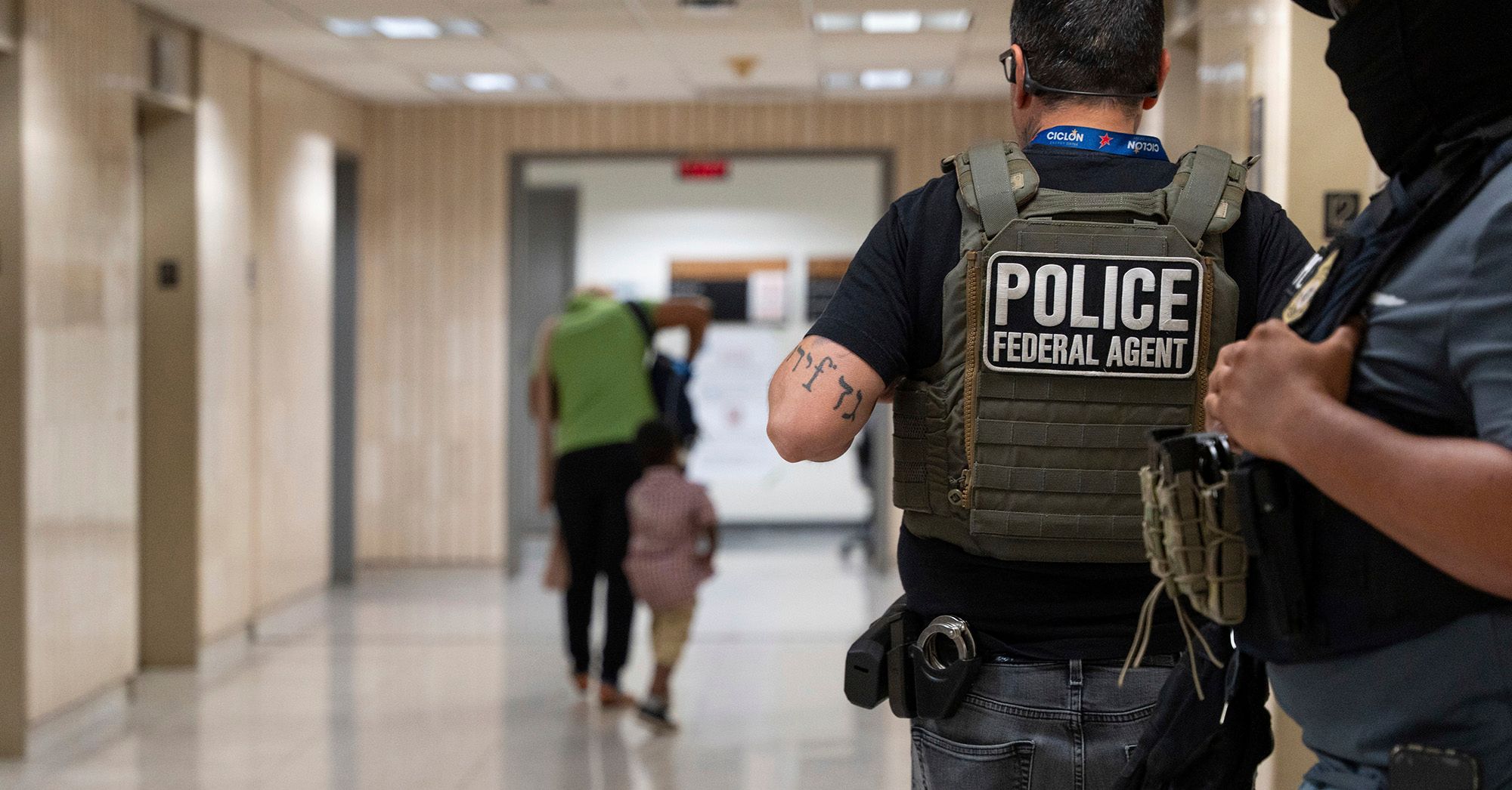 A photo shows the back of a light-skinned officer wearing glasses, a black T-shirt, gray jeans and a vest with the words "police federal agent." One police officer stands next to them. A person and a child walk down a hallway standing in the background.  