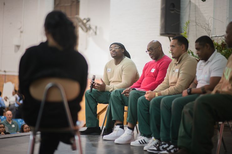 A Black man wearing glasses, dark green prison pants and a tan sweater is holding a microphone as he asks a question to a woman in the foreground, who is sitting on a chair with her back turned to the camera. Alongside the man sit four other men in the same green prison pants and a variety of shirts. Members of the audience look on from their blue-colored seats.  