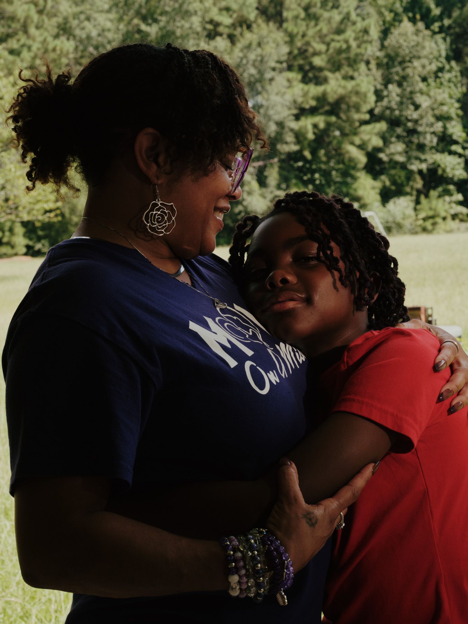 Belinda Gray, a Black woman wearing a purple T-shirt, embraces her grandson, a Black boy wearing a red T-shirt. Trees and grass are visible in the background.