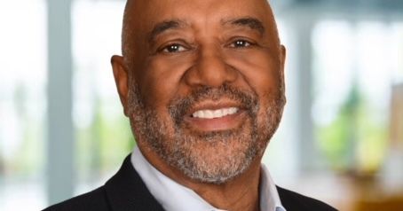 A man with dark-toned skin wears a black suit jacket and white dress shirt.  