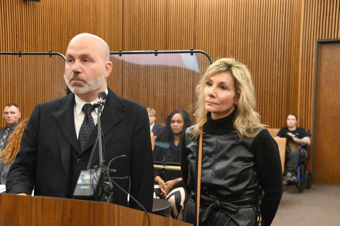 Former Cuyahog County Domestic Relations Judge, Leslie Ann Celebrezze, a White woman with blonde hair, wears a black top and stands behind a podium next to her defense attorney, Ian Friedman, a White man with a beard and wearing a black suit. 