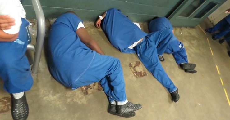 Three men in blue uniforms sleep on the floor, as one sits on a chair nearby.  