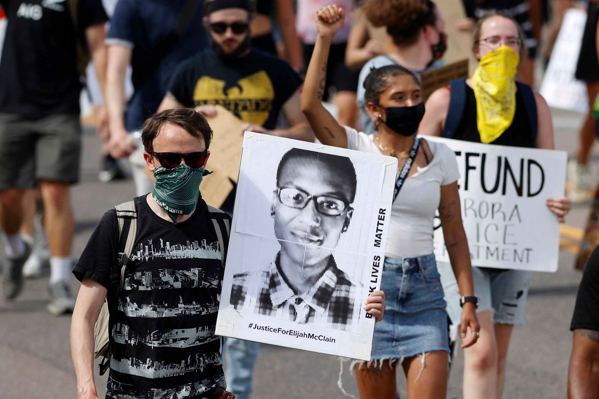 A White person, wearing sunglasses, a green bandana mask and black shirt, holds a poster with the black-and-white image of Elijah McClain, a Black man wearing glasses and a plaid button-up shirt. The poster also reads, "Black Lives Matter. #JusticeForElijahMcClain." There are more protesters marching in the background. 
