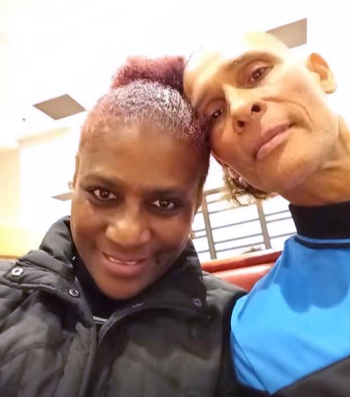A photo shows a woman with dark-toned skin, left, wearing a black jacket, next to a man with medium-toned skin wearing a blue and black top, leaning his head against hers. 