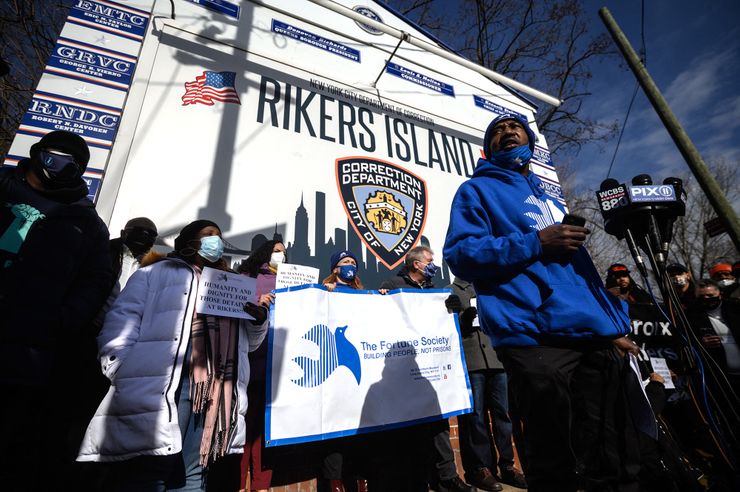 A Black man in a blue sweater, blue mask and black pants speaks into several mics from different TV news stations. A group of protesters stand in front of the Rikers Island jail complex entrance in the background, holding signs that read: "The Fortune Society: Building People, Not Prisons" and "Humanity and Dignity for Those Detained at Rikers!!!"