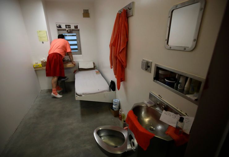 A man in an orange uniform looks out of a small window in his solitary confinement cell, which contains a narrow bed, a sink and attached toilet, and a small desk and chair. 