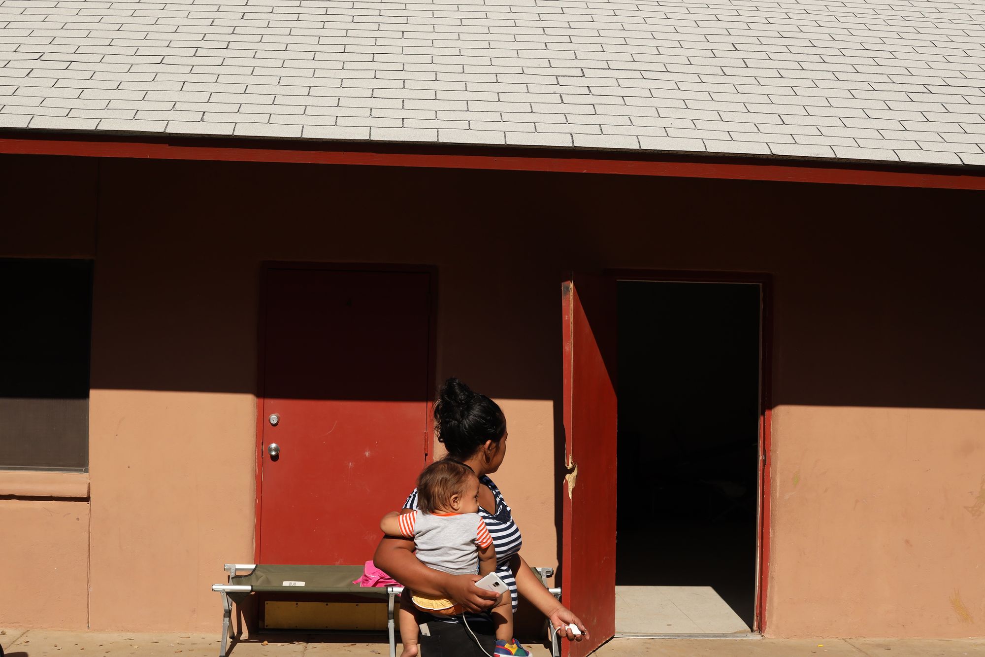 A photo shows a woman with medium skin tone and black hair with her head turned toward an open red door. She holds an infant in one arm.