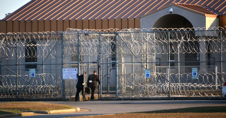 Two individuals in black suits walk through the entrance of a barbed wire fence. A tan prison is behind the fence. 

