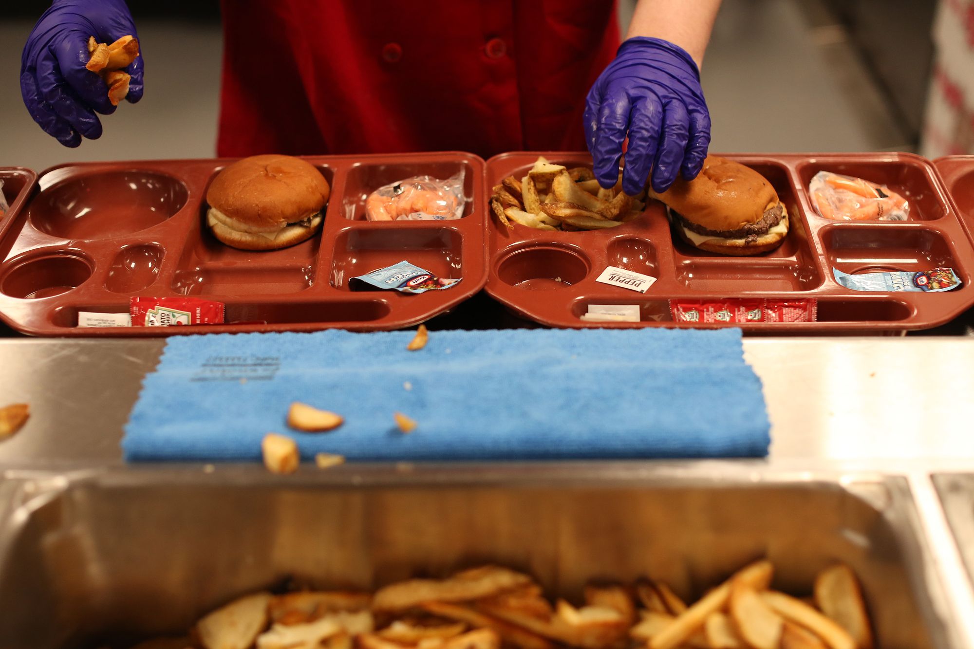 An incarcerated person wearing food preparation gloves adds French fries to a food tray that contains a hamburger and a small bag of baby carrots.  