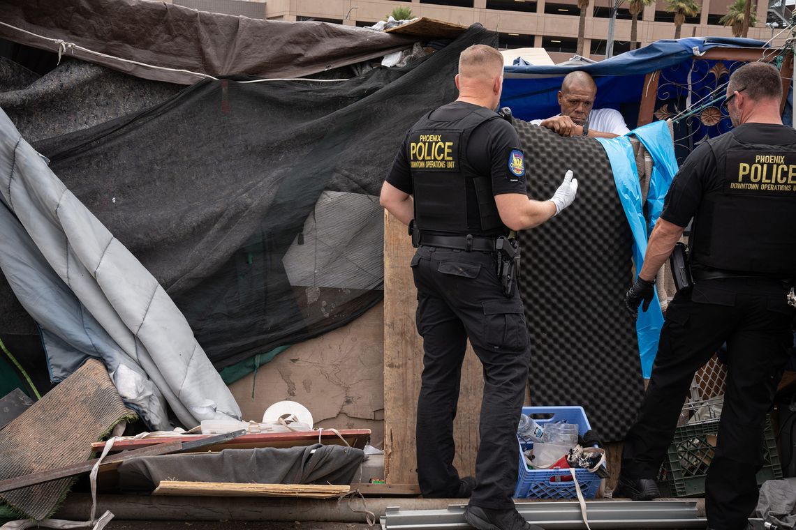 Two police officers, wearing gloves and protective vests with the words “Phoenix police, downtown operations unit” on the back, talk to a Black man at a homeless encampment. 