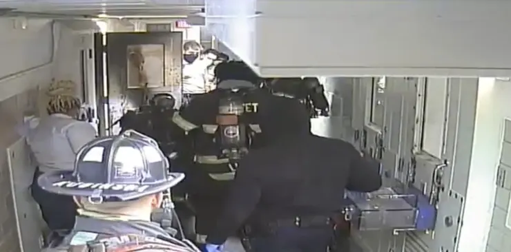 A video still shows firefighters standing in a prison hallway. A cell door with black soot is open on the left. 