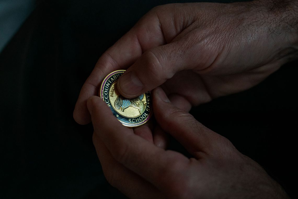 A close-up photo of a White man’s hands holding a large metal coin. 