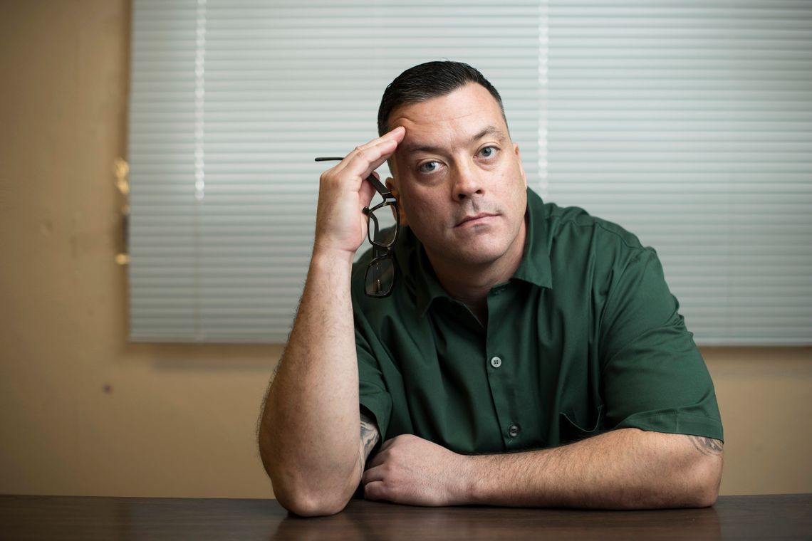 John J. Lennon, a White man with short brown hair, wears a green uniform while holding his glasses in his right hand.  He sits in front of a window that has blinds covering it.