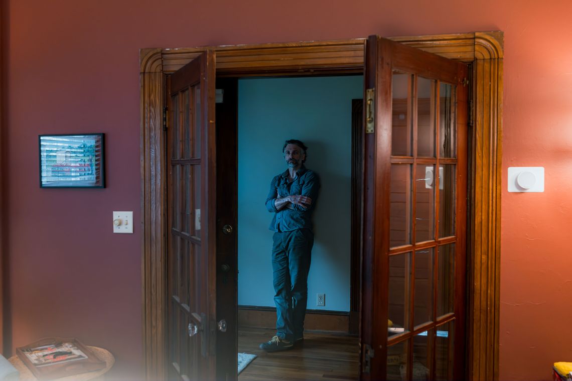 A photo of a middle-aged White man with slicked-back dark hair posing for a portrait and looking toward the camera, through two open doors inside a home. 