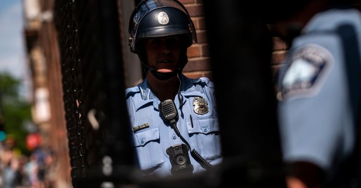 A photograph shows two police officers standing next to a gate on a street. One officer is wearing a helmet and holds a wooden baton. 