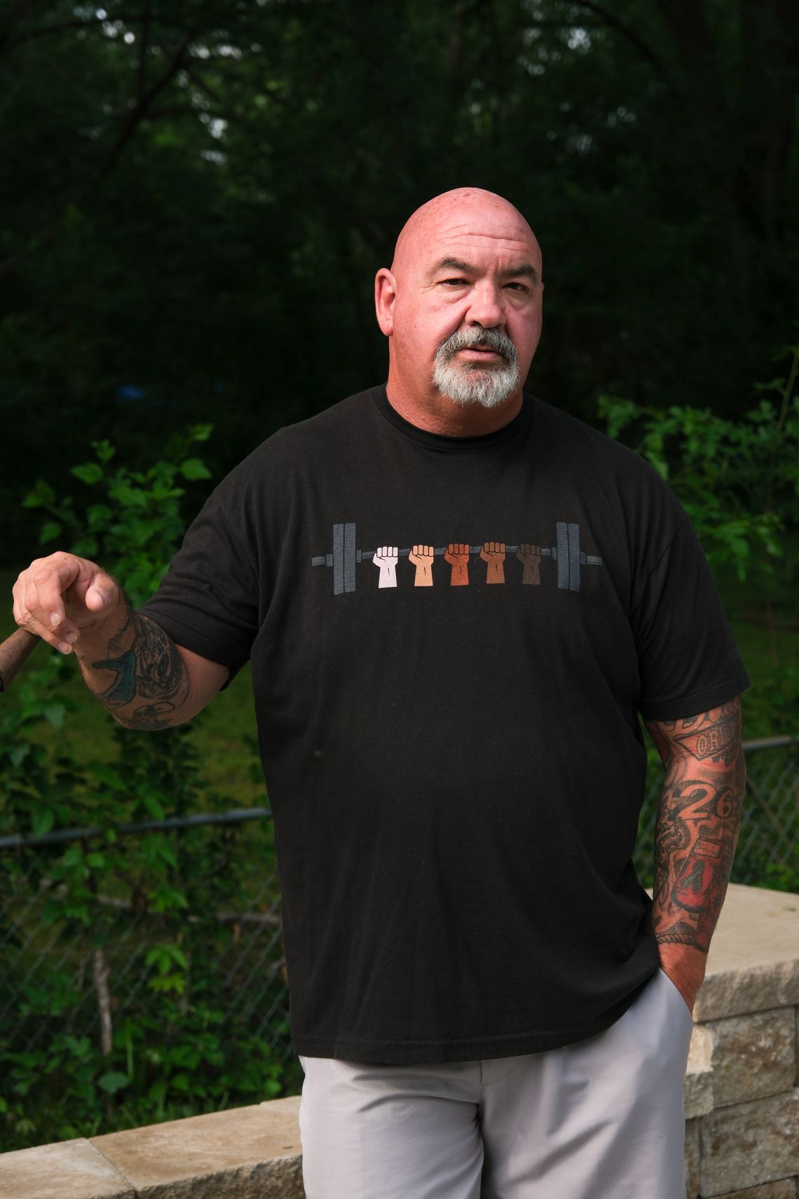 A White, bald man with gray facial hair and wearing a black T-shirt stands against green foliage.