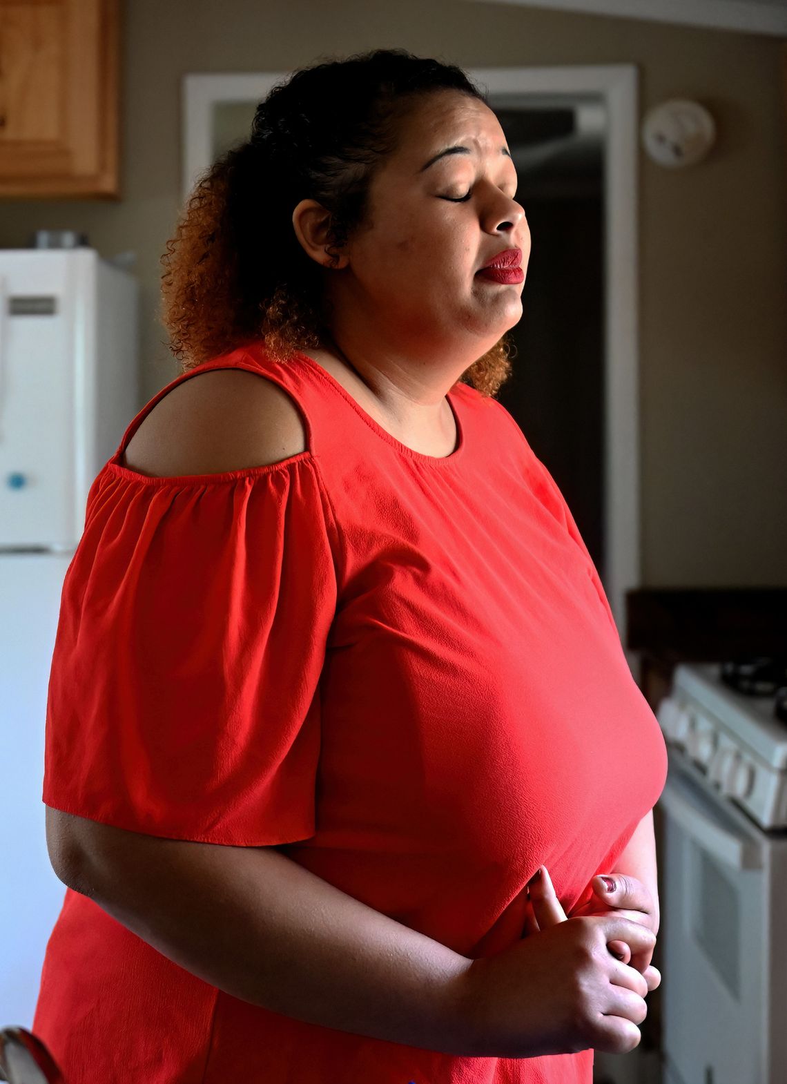 A Black woman, wearing a red blouse and red lipstick, has her eyes closed and her hands clasped together while she stands in a kitchen. 