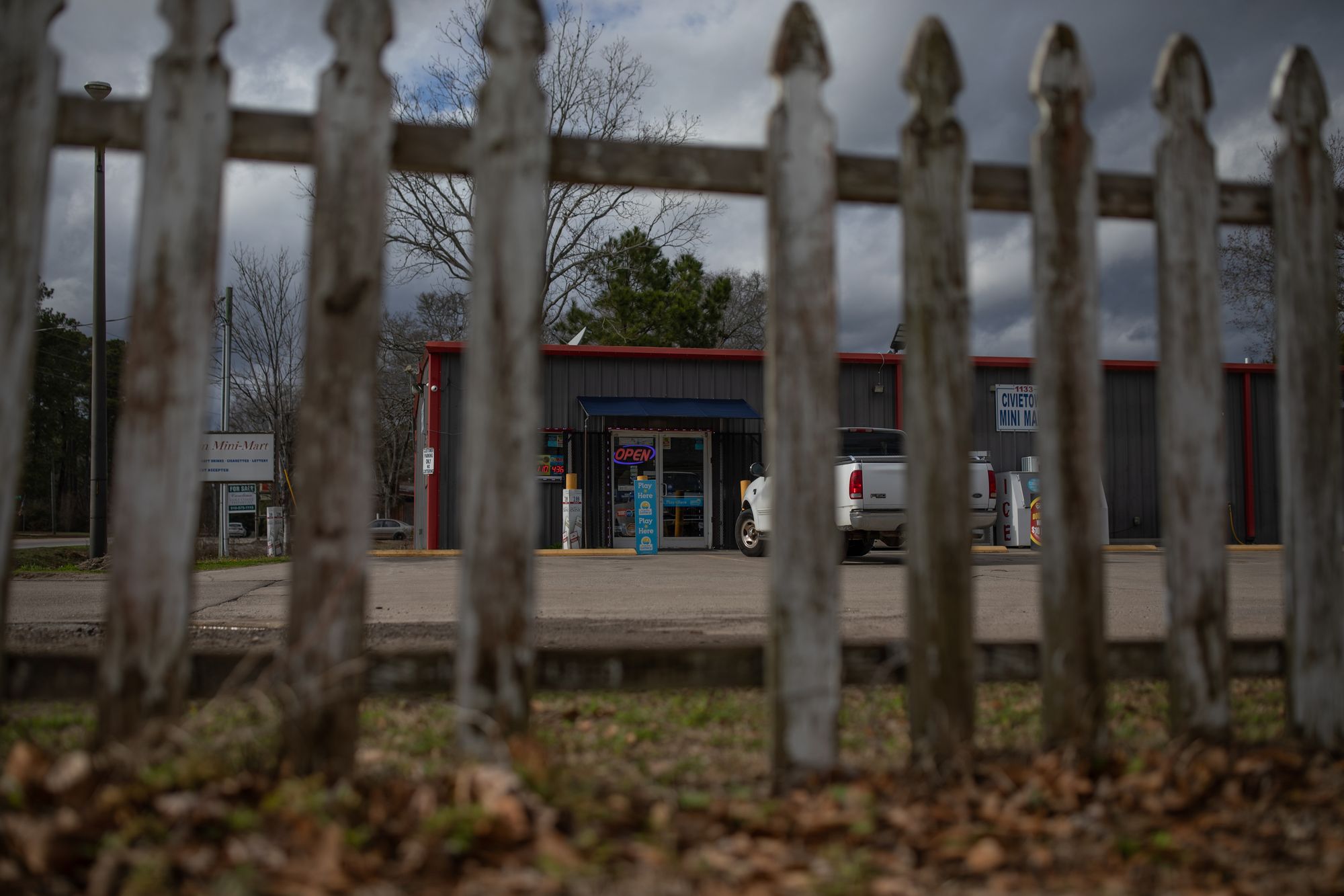A photo shows a mini-mart behind a wooden fence that has a slat missing in the middle. A white truck is parked outside the mini mart. 