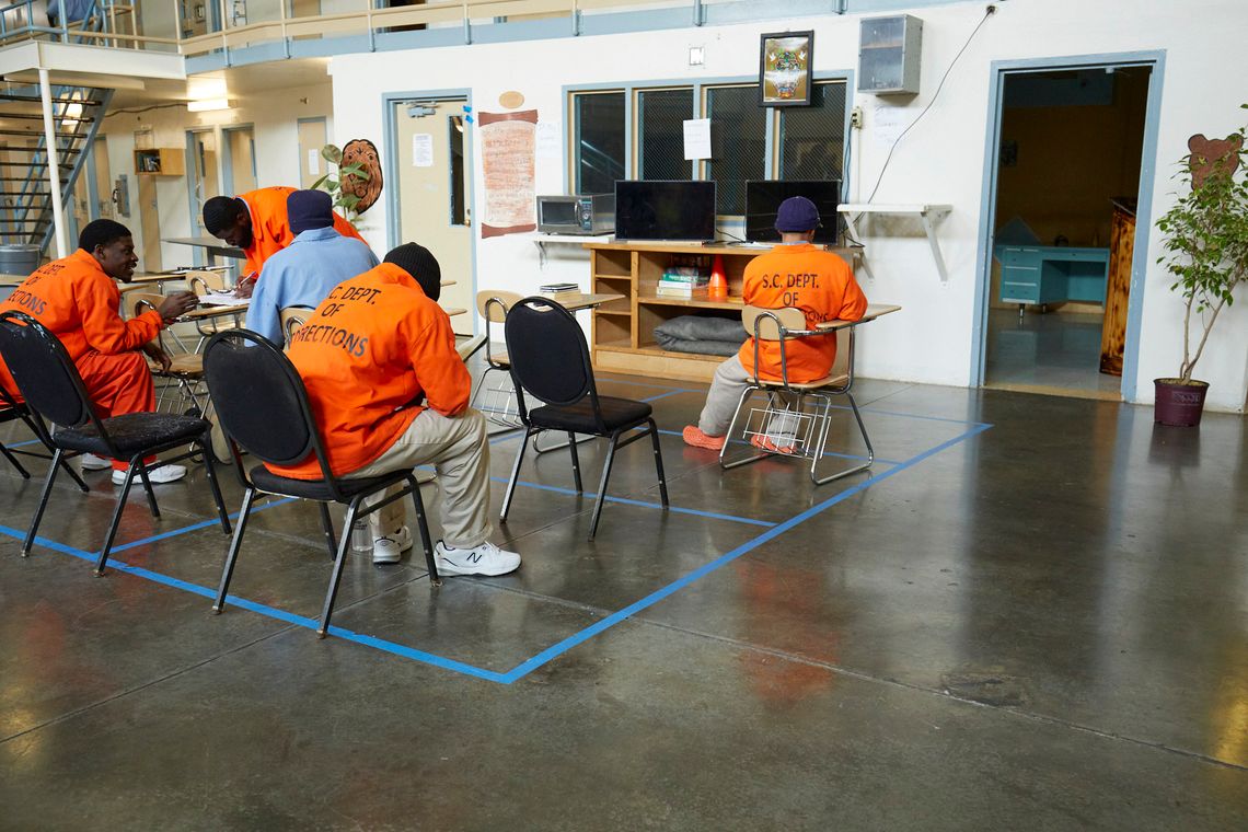 Four students wear orange coats reading “S.C. Dept. of Corrections” on the back, and one wears a light blue button-down shirt while sitting in a classroom setting.