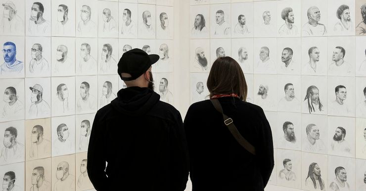 Two people, wearing all black clothing, stand and view more than a hundred portraits of incarcerated men on display, on an art gallery wall. 