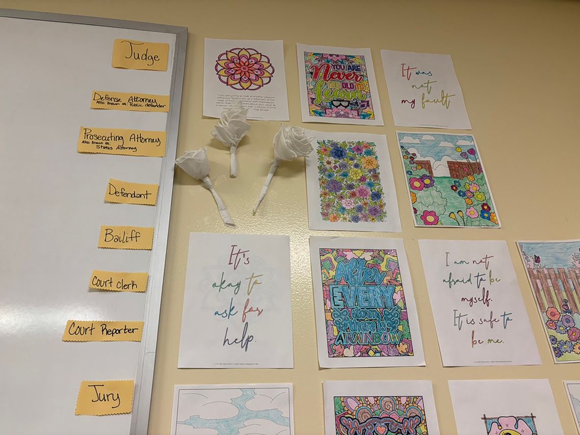 A photo shows multiple pieces of paper and paper flowers taped to a wall. To their left, several yellow index cards are taped down the side of a whiteboard, with terms written on them such as “judge,” “defense attorney,” "”prosecuting attorney,” “defendant,” “bailiff,” “court clerk,” “court reporter” and “jury.”