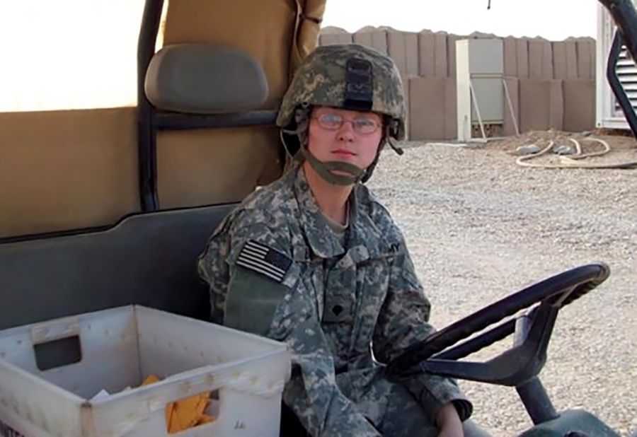 A White woman, wearing glasses, Army Reserve uniform and helmet, sits in a cart. A white mail dropbox filled with manila envelopes is next to her. 