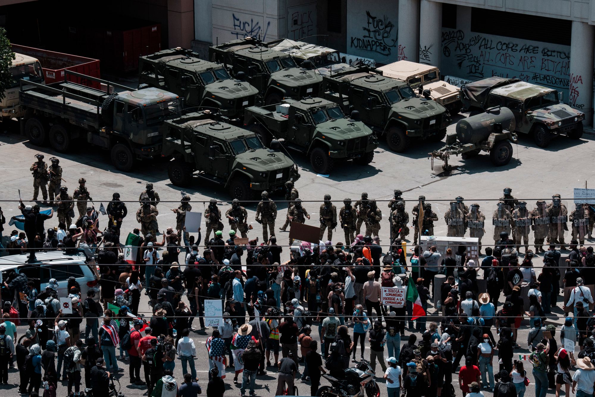 A large group of protesters stand with signs in the lower half of the photo, facing green military-grade vehicles.  